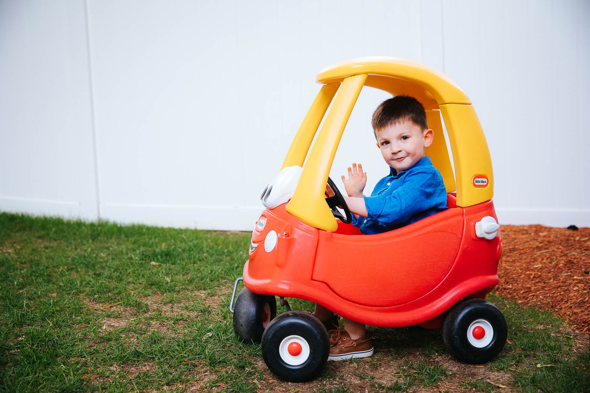 4 year old boy honks horn as he rides through his backyard in a red Fischer Price car
