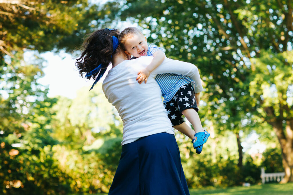 Little boy laughing in his mother's arms as she spins him around, green leaves behind him.