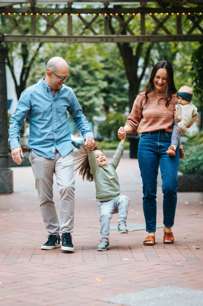 Father and mother swinging their toddler son between them as they walk through the financial district of Boston, their newborn daughter resting on the mother's right hip.
