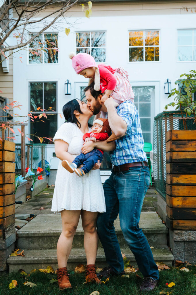 Family of 4 in front of their Cambridge home, mother and father kissing, holding infant between them as toddler daughter sits on top of father's shoulders