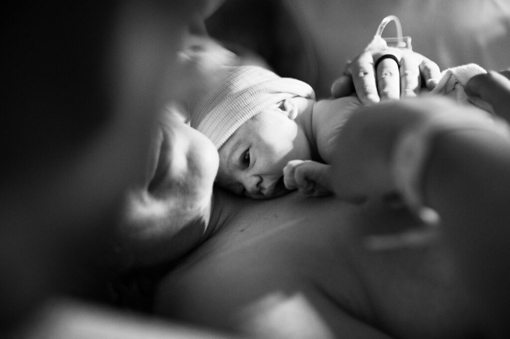 Black and white image of a baby just after being born in the hospital. He clings to his mother's bare chest as she smiles.