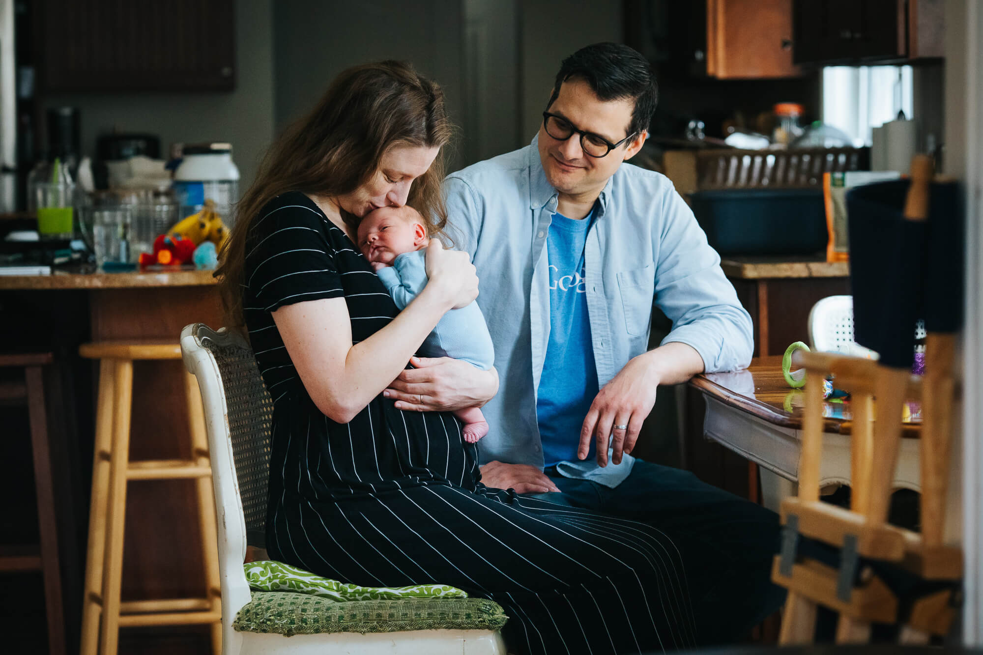 Young mother sits on a wooden chair in a dining room surrounded by bottles and breastfeeding gear. She holds her newborn son and nuzzles the top of his head, smiling. Father sits next to her and smiles while gazing at her face.