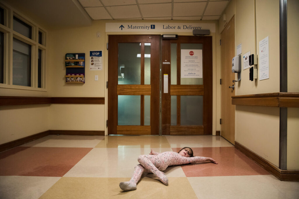 A little girl in her pajamas rolling around on the floor of a hospital waiting room as her little sister is born