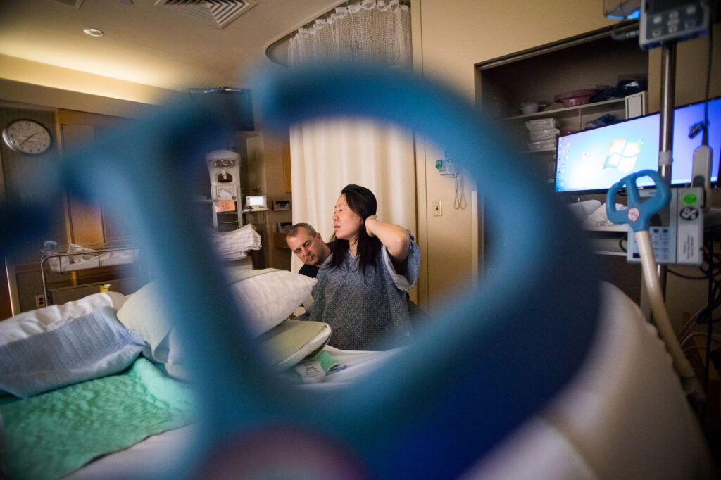 View through bed stirrups of male partner holding birthing partner's waist as she labors, both of them smiling at each other