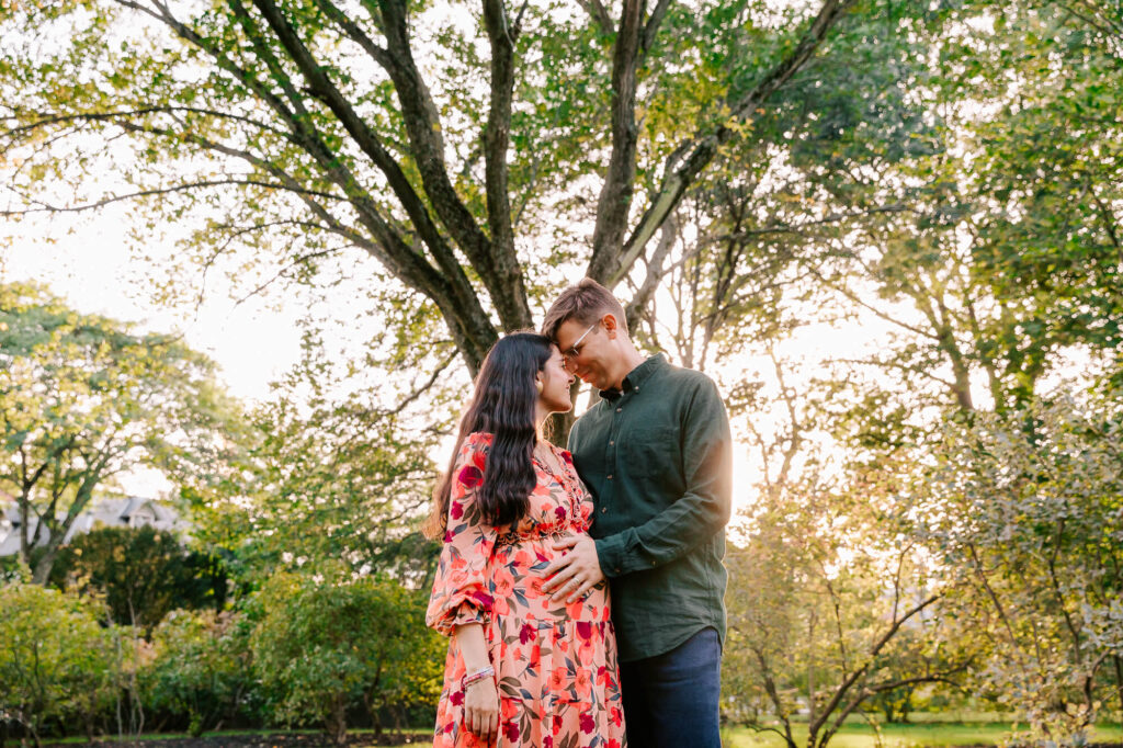 Pregnant couple in an embrace under a large maple tree, with the sun shining through the branches.