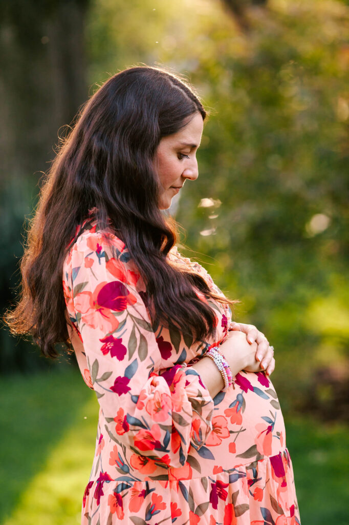 Pregnant woman rests her hands on her belly and looks down. The sun shines in her dark hair.