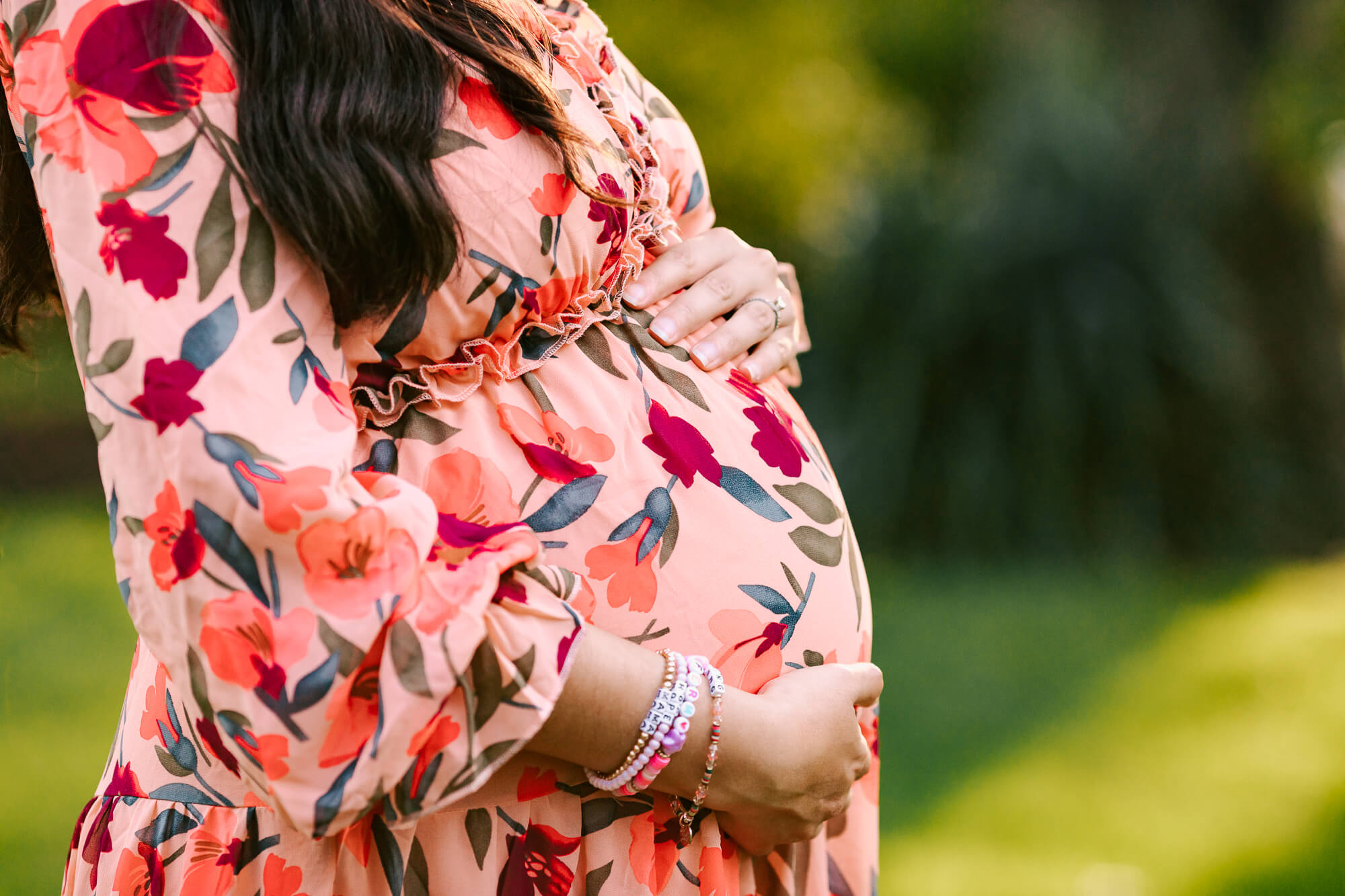 Image of a woman's torso only, with her hands gently framing her stomach. She is about 7 months pregnant and is wearing a pink flowered dress, with a green lawn behind her.