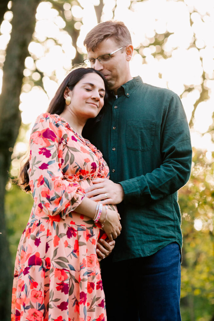 Husband kisses the top of his pregnant wife's head. She is wearing a pink flowered sundress and has long dark hair.