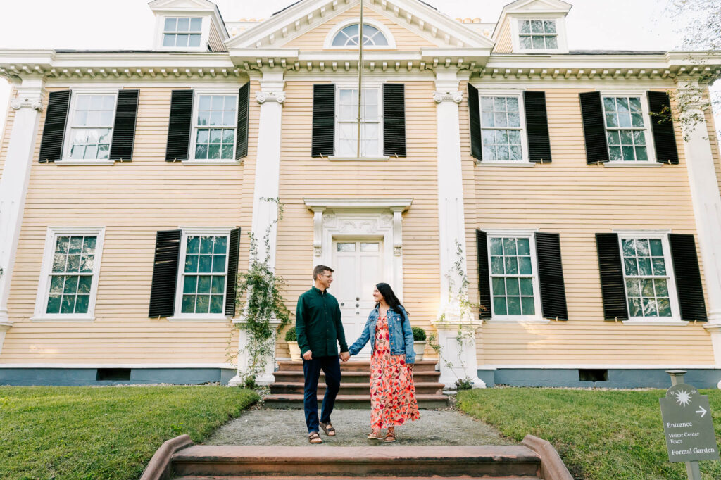 Man and woman laugh together as they walk down a set of stairs in front of a historical home in Cambridge. She is pregnant and they are both smiling.