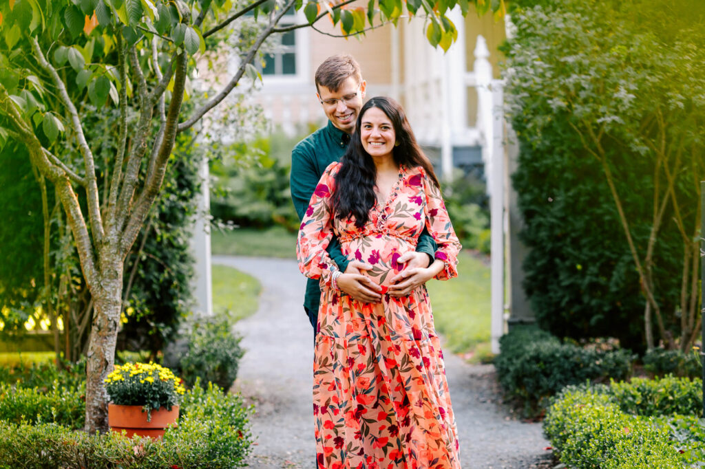 Pregnant couple stands at a garden gate. She is smiling and looking at the camera, he is behind her and looking down as he holds her stomach.