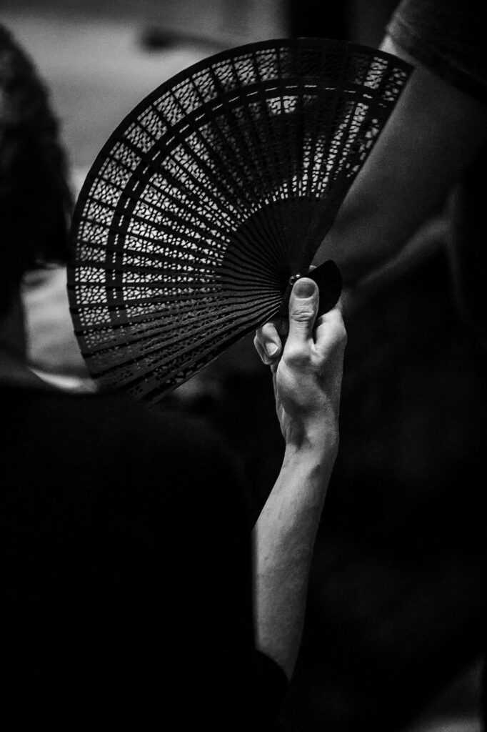 Black and white image of the back of a woman's hand holding a plastic fan; through its holes we see the edge of a bathtub