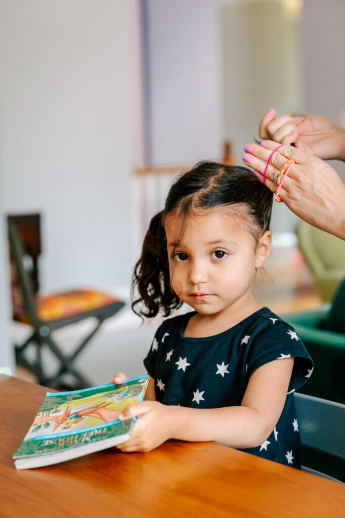 Little girl in black dress with white polka dots sits in dining room chair as her mother pulls one section of hair back into a pigtail.