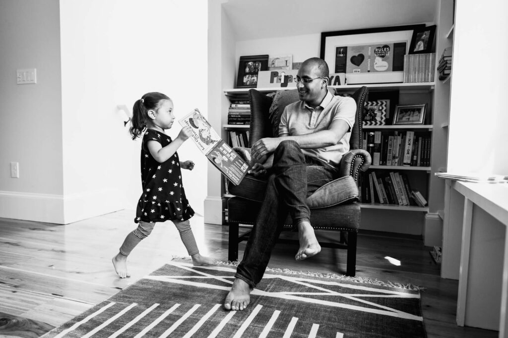 Black and white image of 3 year old girl running to her father with a picture book. He sits in an armchair by the window and smiles at her.