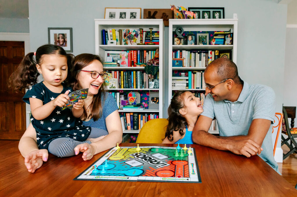 3 year old girl with pigtails sits in her mother's lap, as 6 year old girl next to them looks up lovingly at father. They're sitting at their dining room table playing the board game "Sorry!"