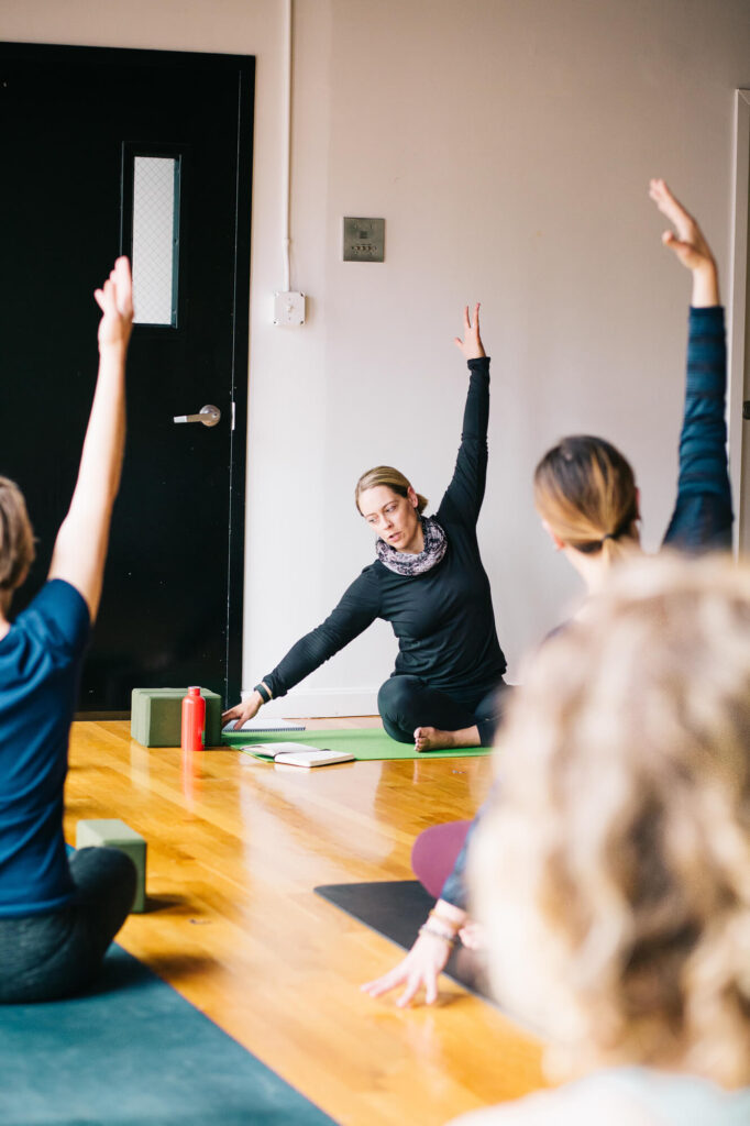 A yoga teacher demonstrates a side stretch at the front of her classroom