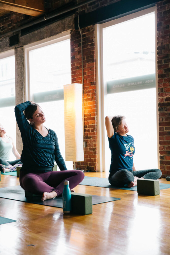 Two students sit on their mats near large windows in a Medford MA yoga studio