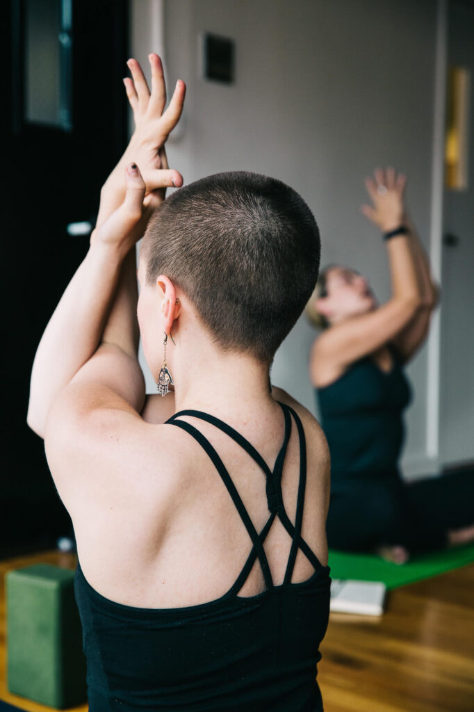 View from the back of a woman practicing a yoga arm bind in a Medford MA yoga studio. She's wearing a strappy black tank top and has buzzed short hair.