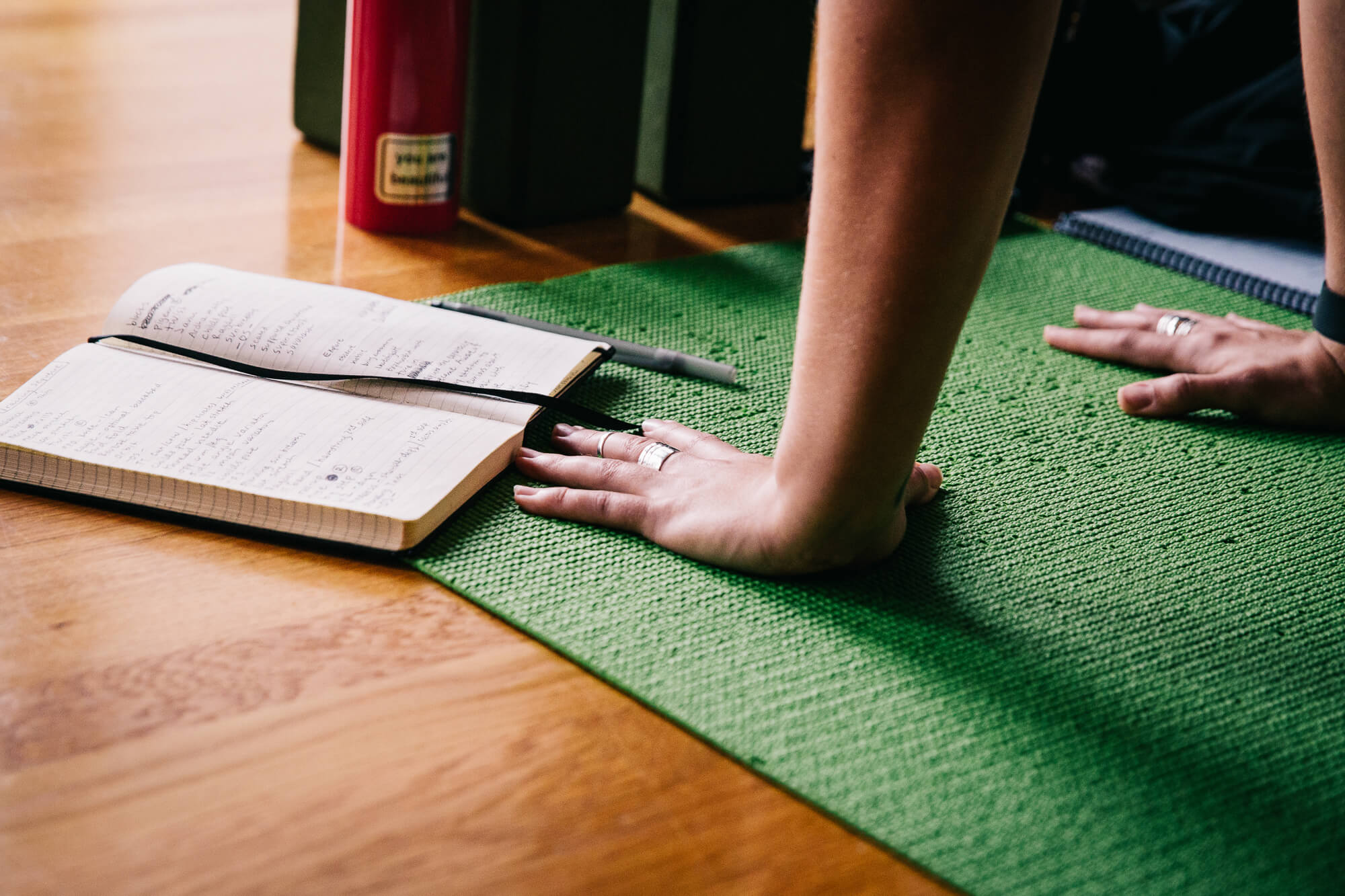 Yoga teacher demonstrates a pose on her mat: we just see her hands planted at the top of a green mat, with an open notebook and water bottle nearby.