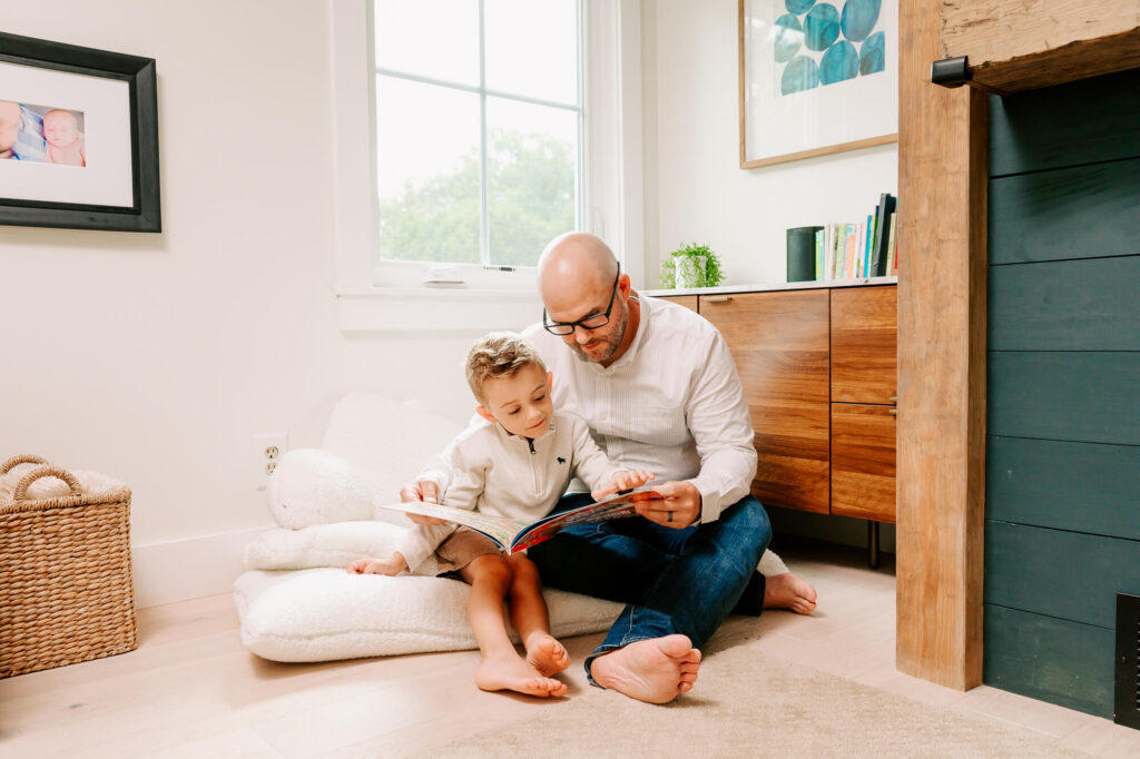 Dad in white dress shirt with shaved head and stubble sits on a cushion under a bright window and reads to his 6 year old son, who cuddles into him.