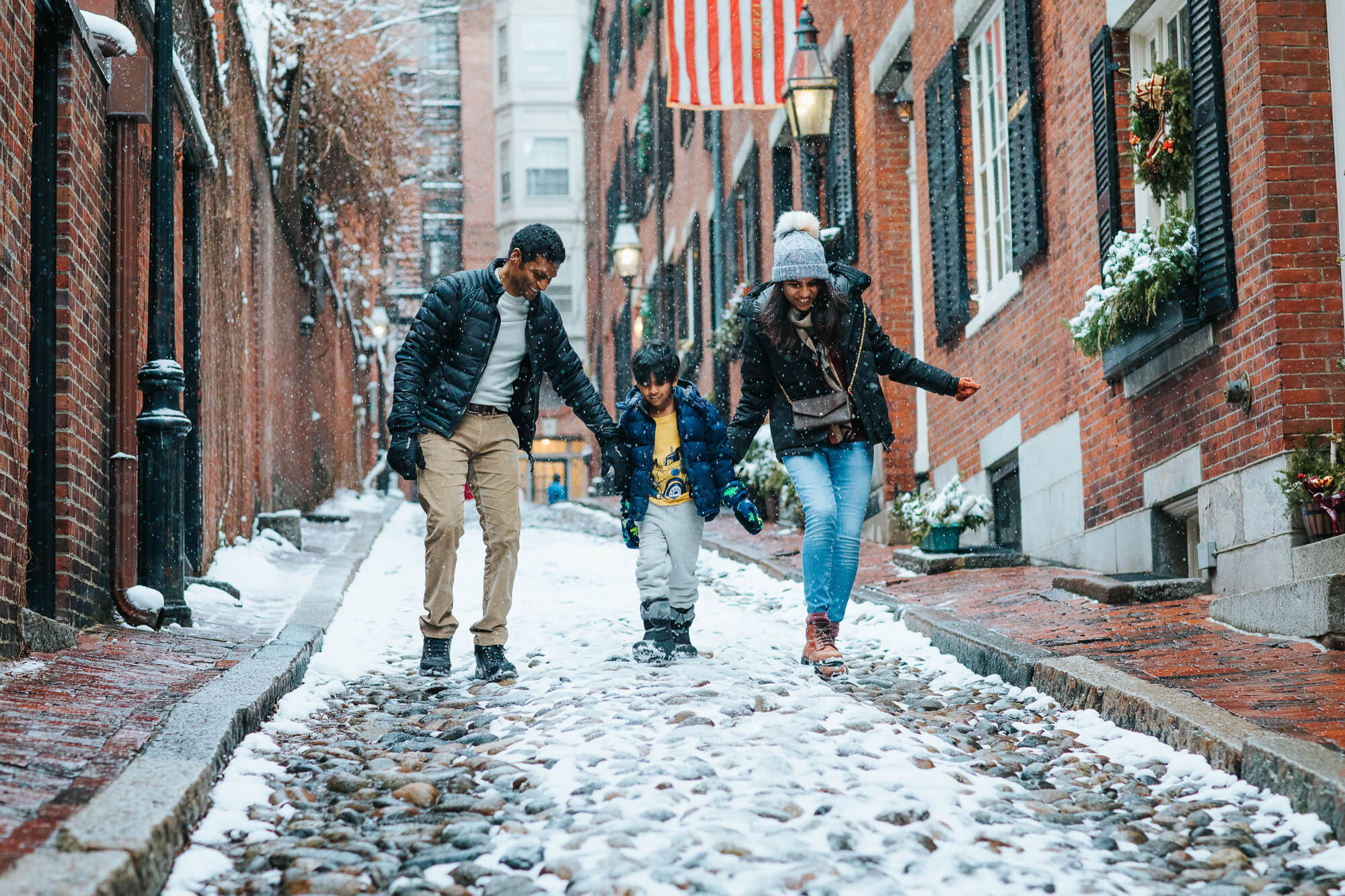 Mother and father hold their smiling 5 year old son's hands and walk down a cobblestone street in Boston's Beacon Hill; they're in winter coats and it's snowing, a light layer of white coats the street.