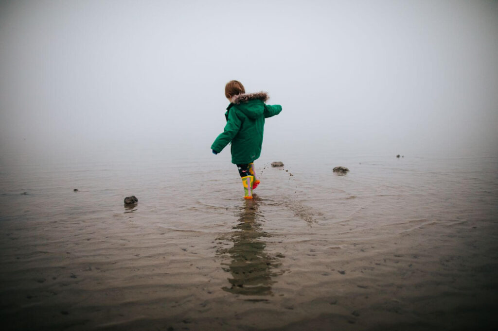 4 year old boy, seen from behind as he splashes along the beach in bright yellow rain boots and a forest green winter coat. There is a wall of white fog ahead of him.