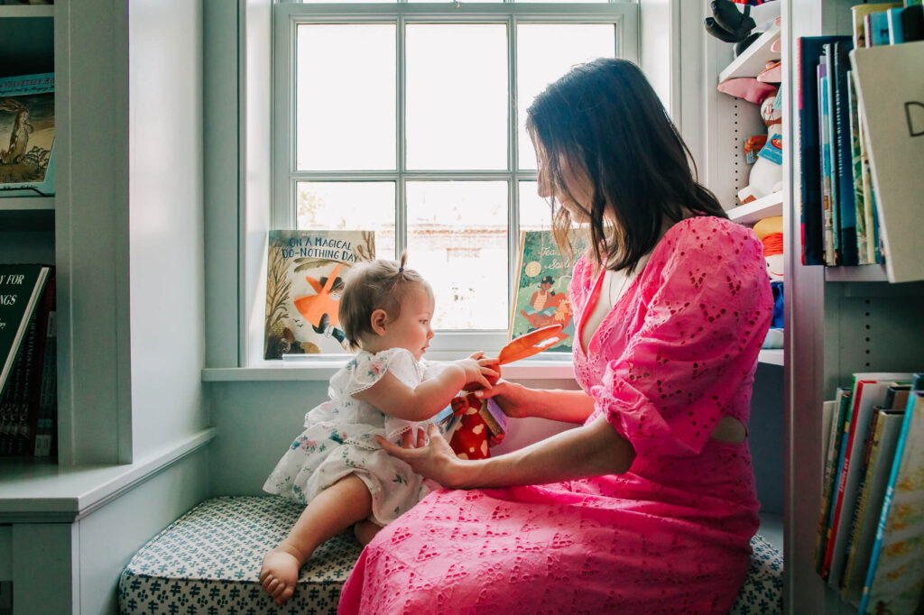 Mother and 1 year old daughter sitting in windowseat of Beacon Hill Books; she is wearing a bright pink dress and smiling as her daughter examines a toy.