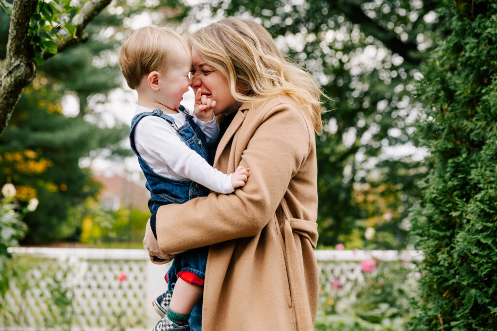 Blonde mother in a tan camel hair coat holds her toddler son close; they are smiling and touching noses, evergreens behind them.
