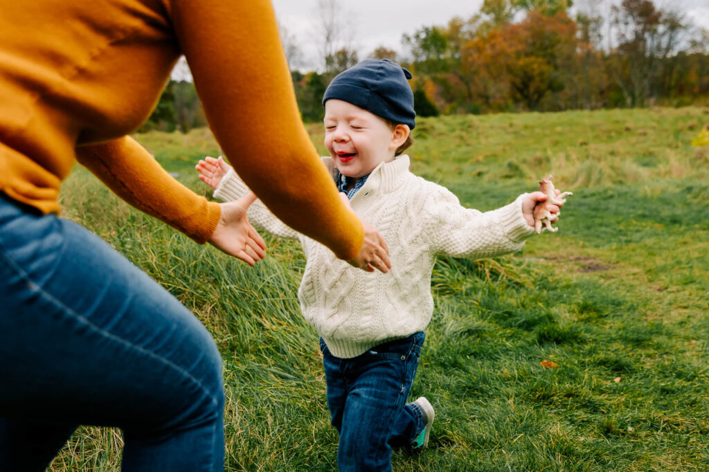 3 year old boy in a blue knit hat and white sweater runs toward his mother's outstretched arms, his eyes closed.
