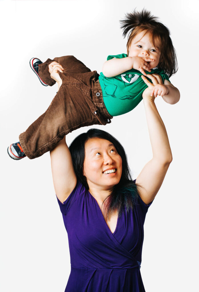 Young dark-haired mother in purple dress holds her laughing toddler over her head. The background is pure white.