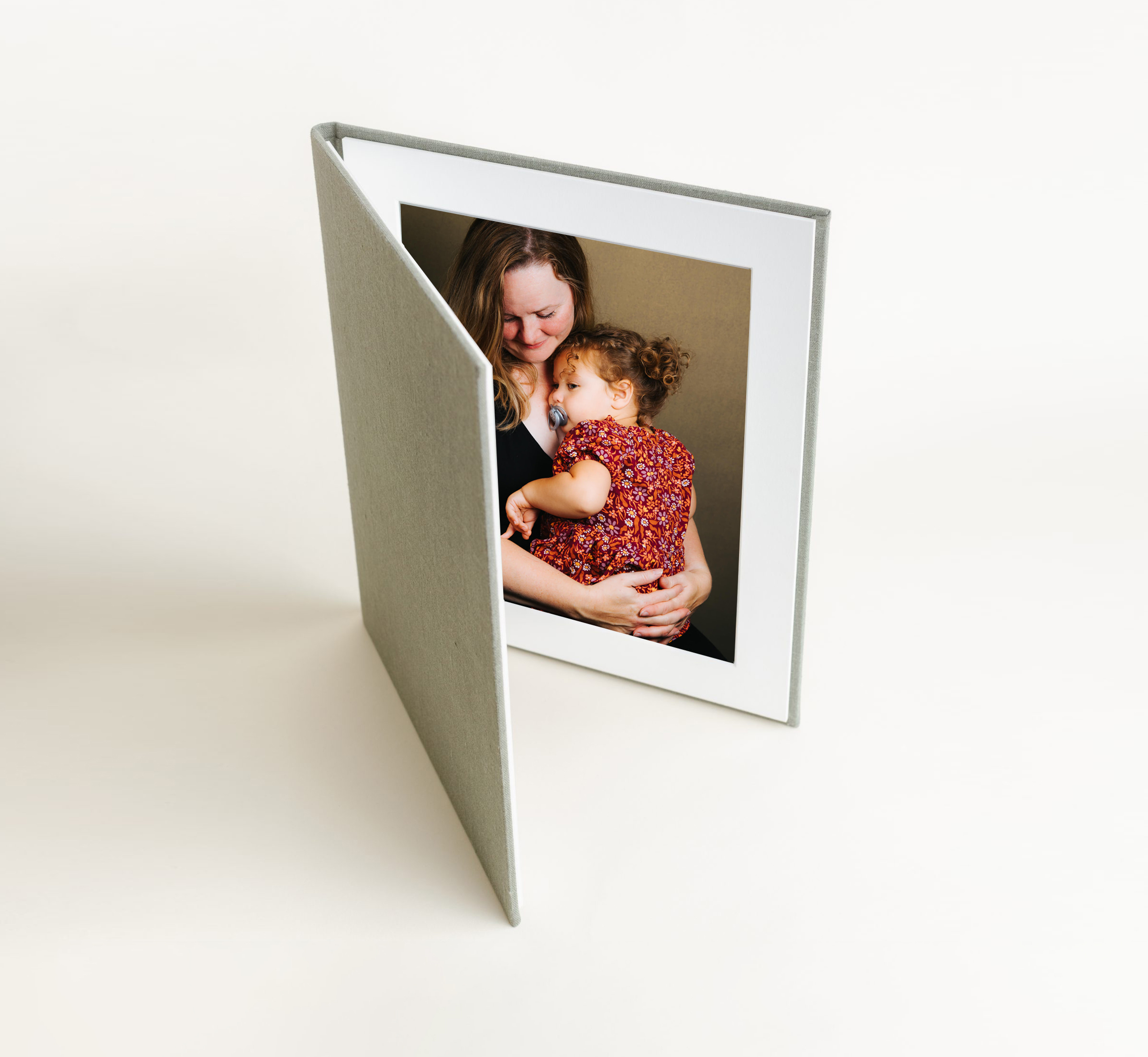 Light green photo folio stands on a shaded cream backdrop. Image inside folio is of a mother holding her snuggled-in toddler daughter, both of them looking down.