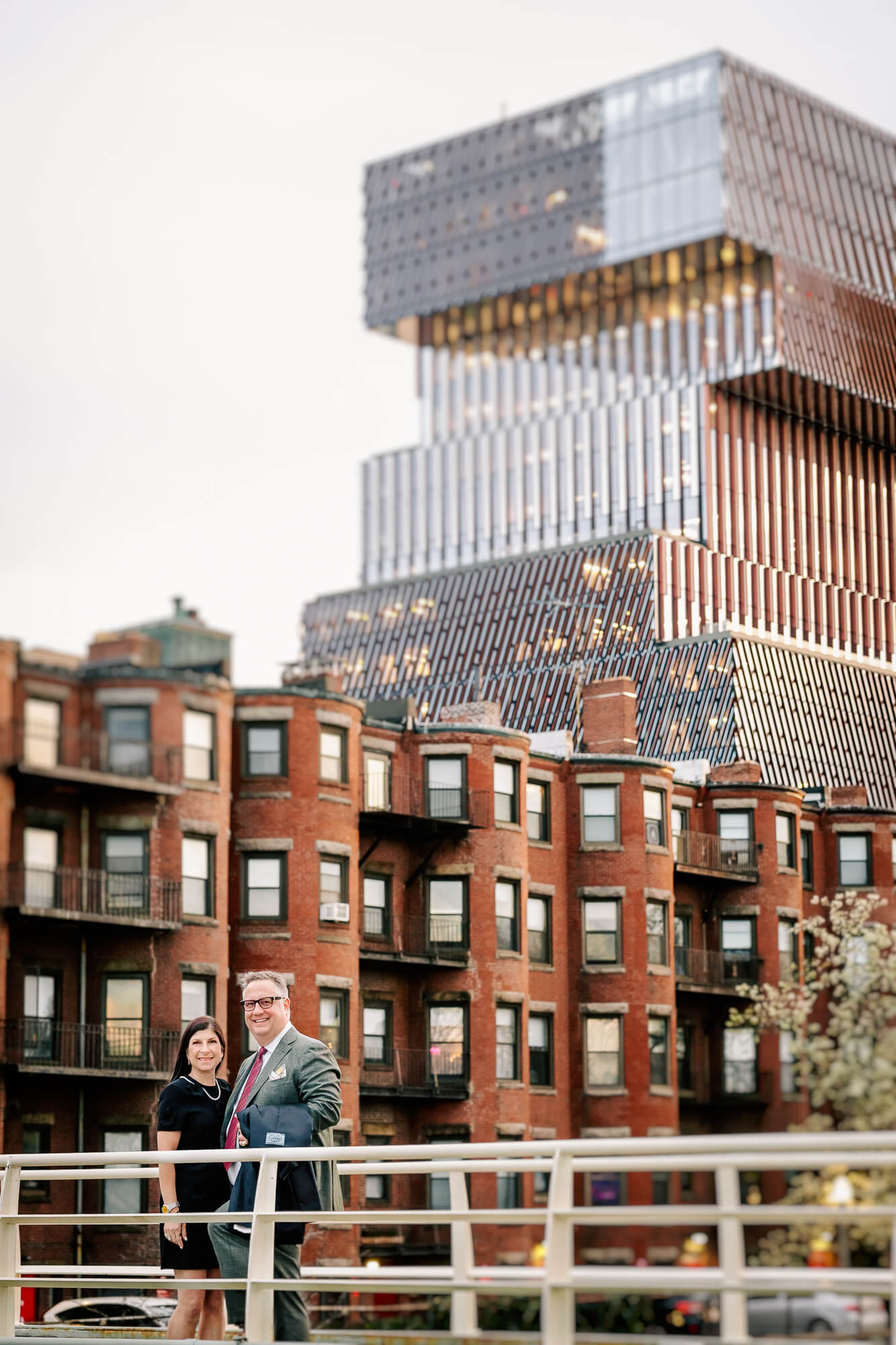 Smiling couple stands on a bridge with brownstones and a large reflective city building behind them