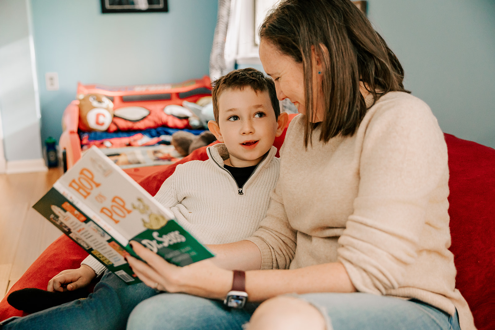 A mom smiles while reading a book to her young son on a bean bag chair in his room