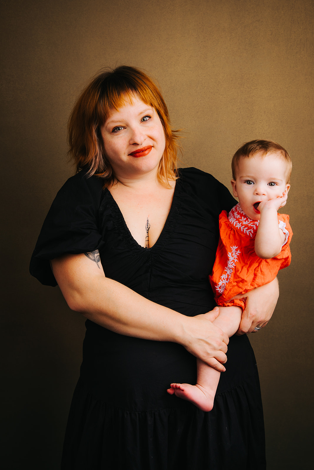 A happy mom stands in a studio in a black dress with her baby in an orange onesie on her hip