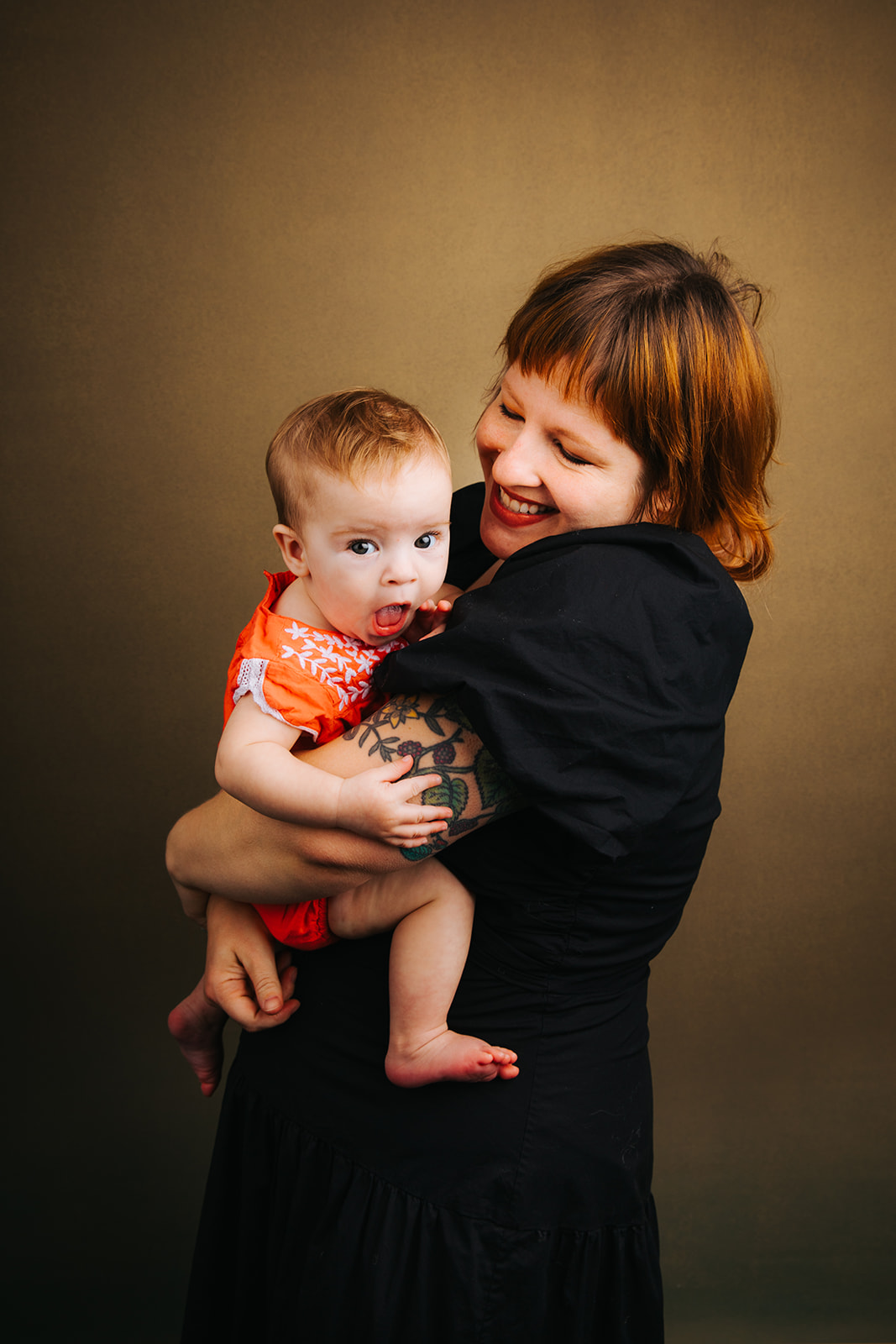 A happy mother hugs and laughs with her baby in her arms in an orange onesie and black dress before some baby music classes in Boston