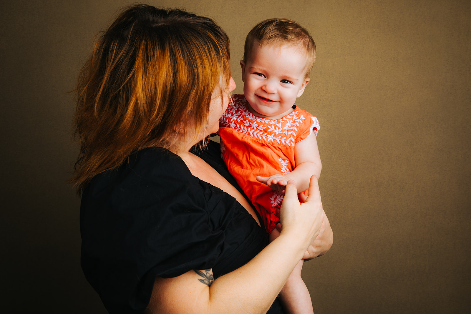 A baby sits in mom's arms in an orange dress in a studio before some baby music classes in Boston