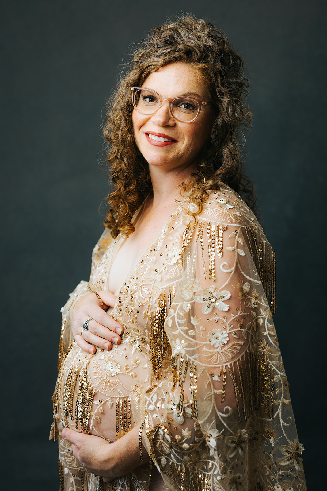 A mother to be with glasses holds her bump and smiles in a studio wearing a gold lace maternity gown