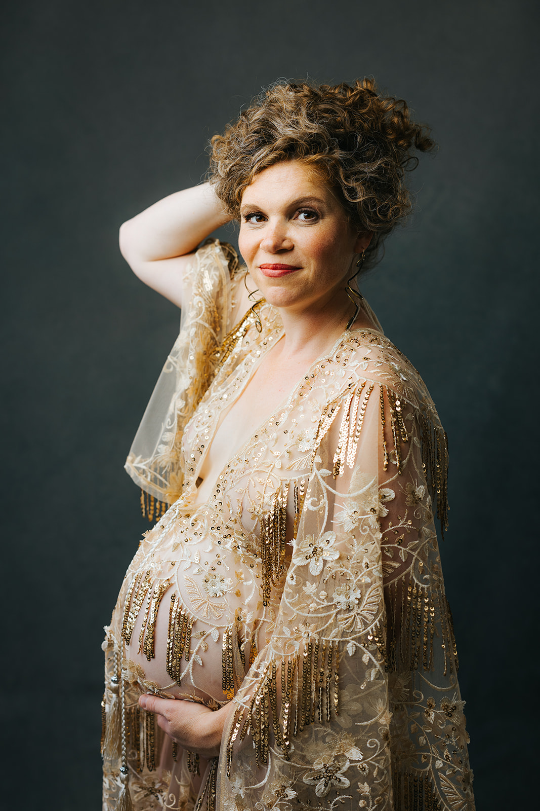 A happy mom to be stands holding her hair up in a gold maternity gown of lace in a studio