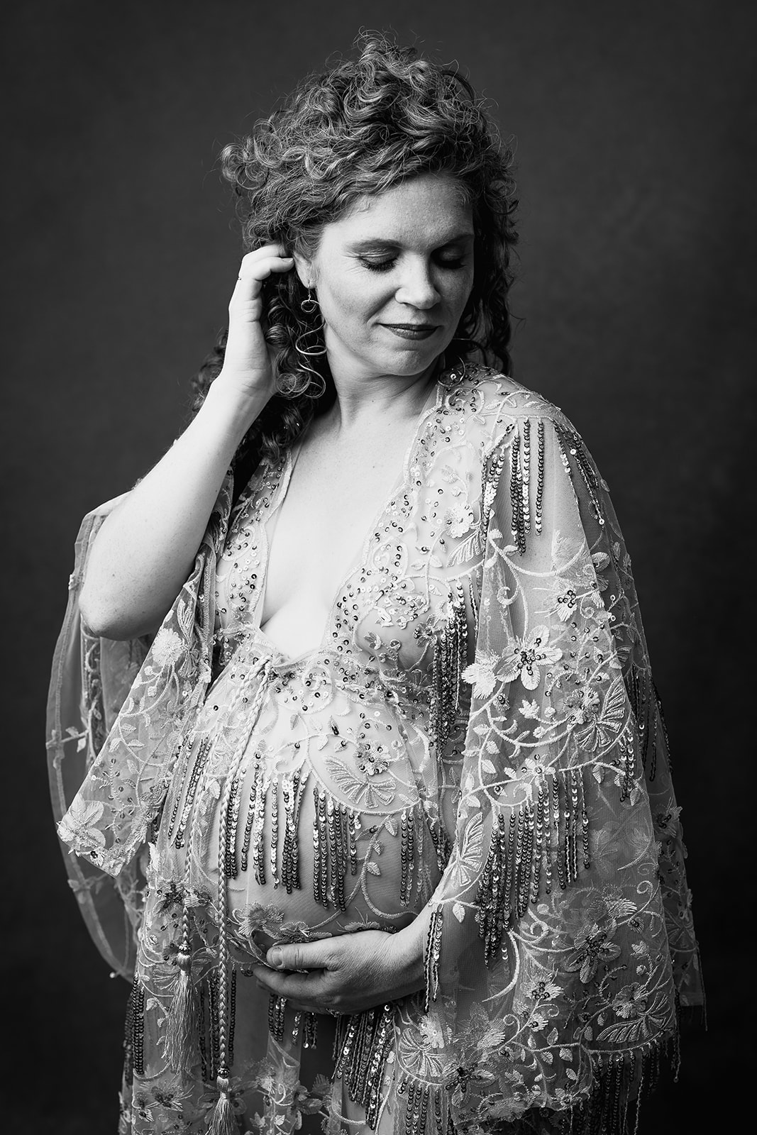 A pregnant woman in a lavish lace maternity gown pulls her hair back in black and white while standing in a studio after visiting baby shower venues in Boston