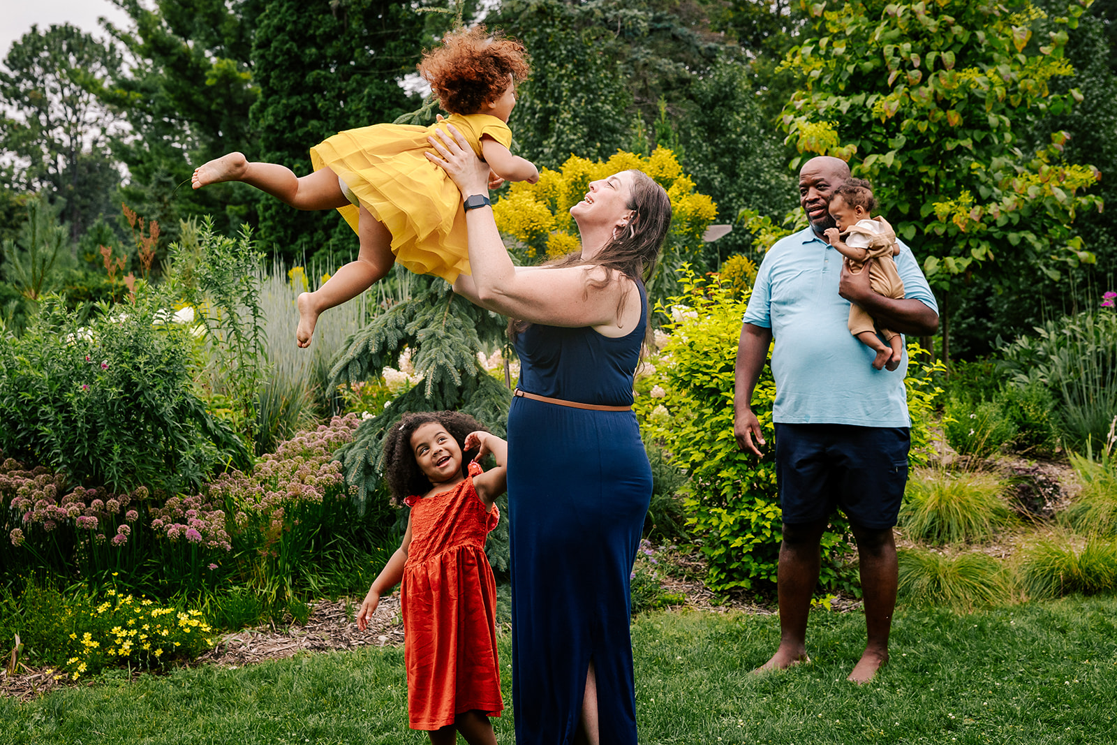 A mom tosses up her toddler daughter in a yellow dress as dad and other 2 daughters look on