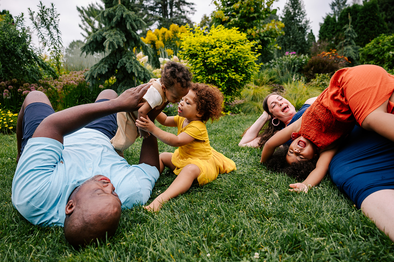 A mom and dad laugh and play with their three toddlers climbing on them in the grass of a garden