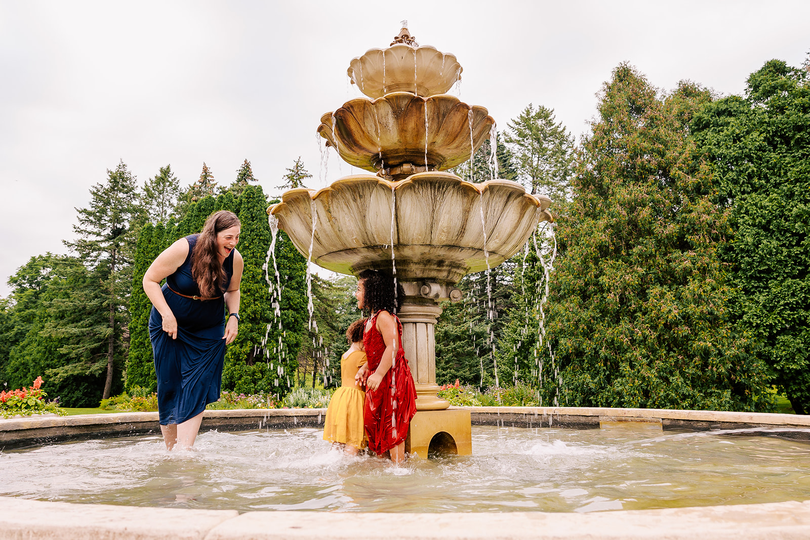 A mom plays with her two toddler daughters in dresses in a fountain in a garden after visiting a Boston daycare