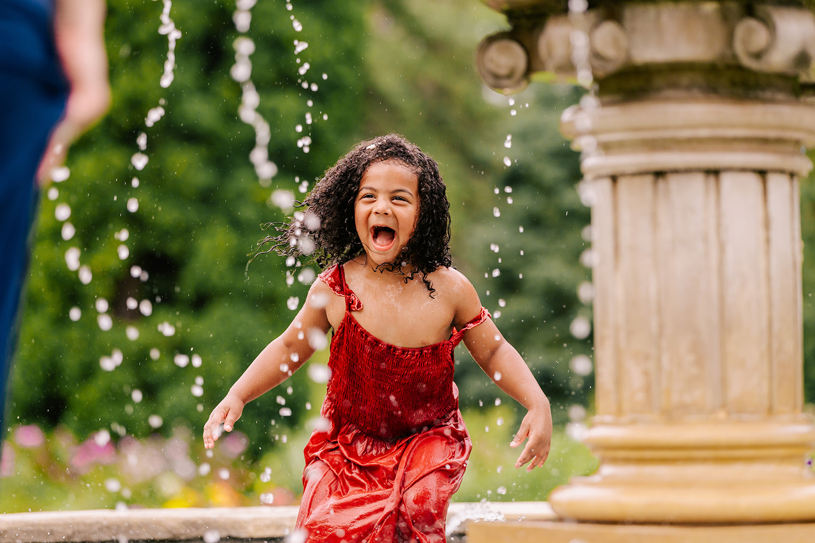 a toddler girl in a red dress laughs while playing in a splash pad after visiting a Boston daycare