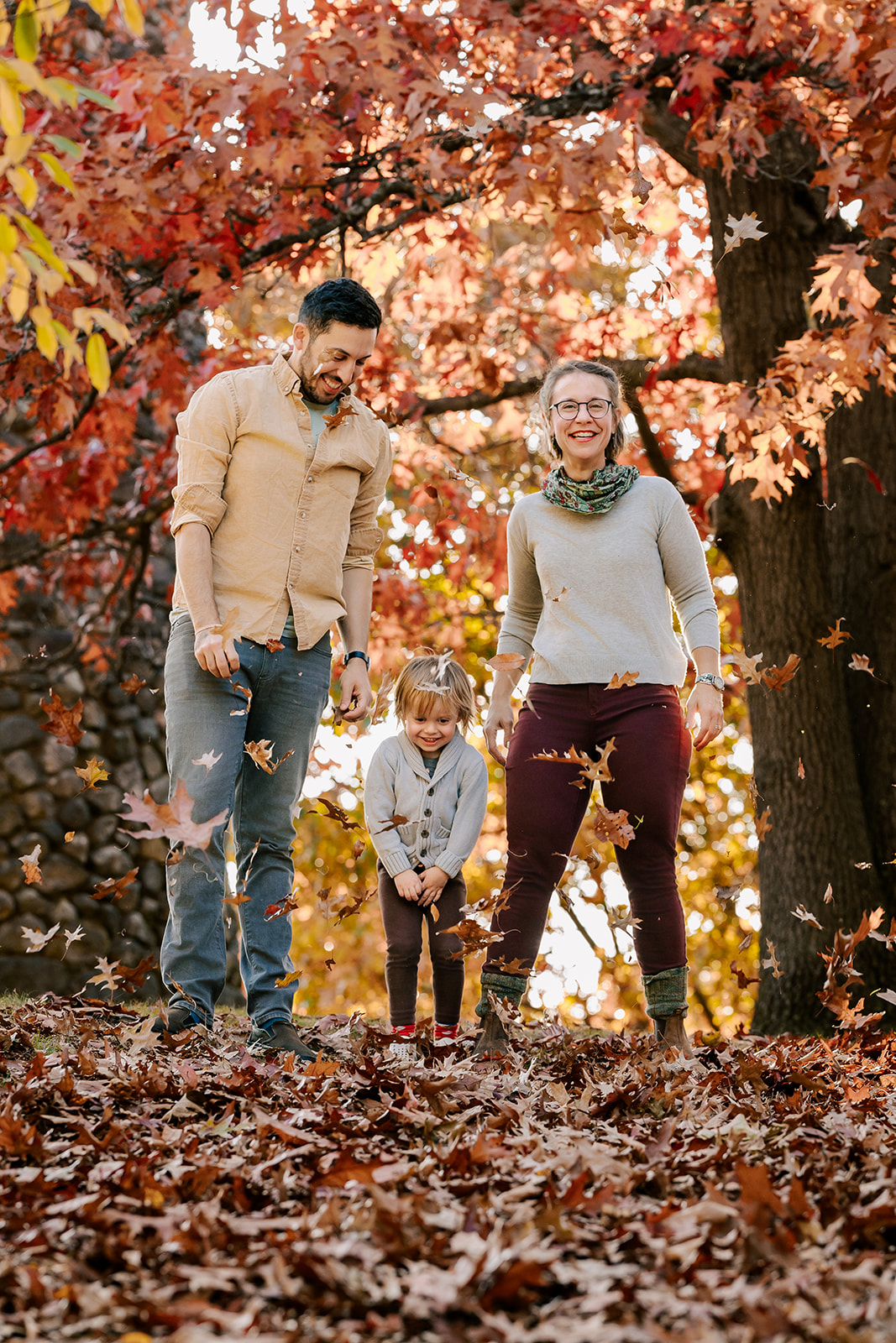 A mom and dad smile and play with a toddler in a park in fall at sunset