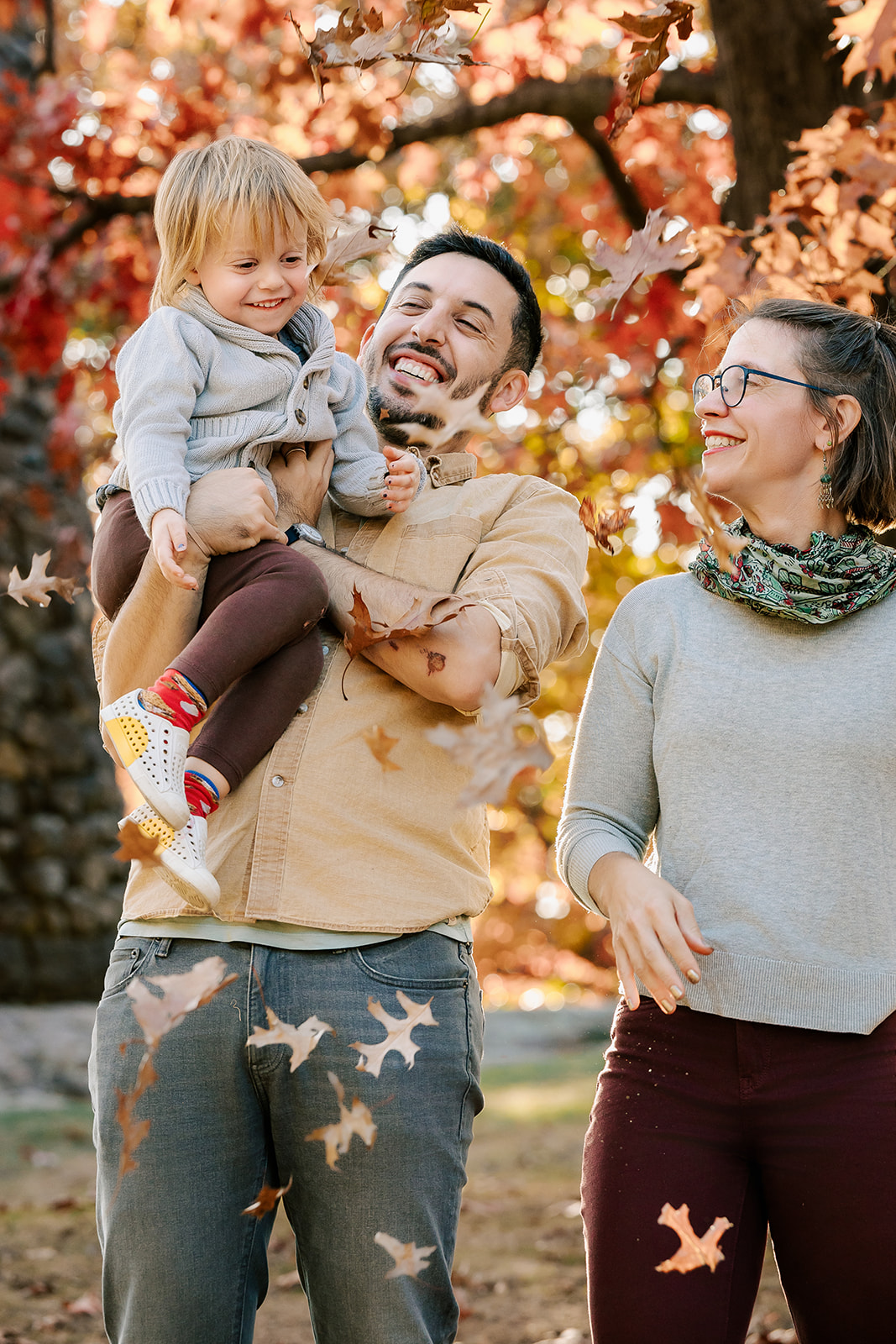 A toddler laughs with mom and dad in a park with leaves falling around them in fall after meeting Boston Nannies