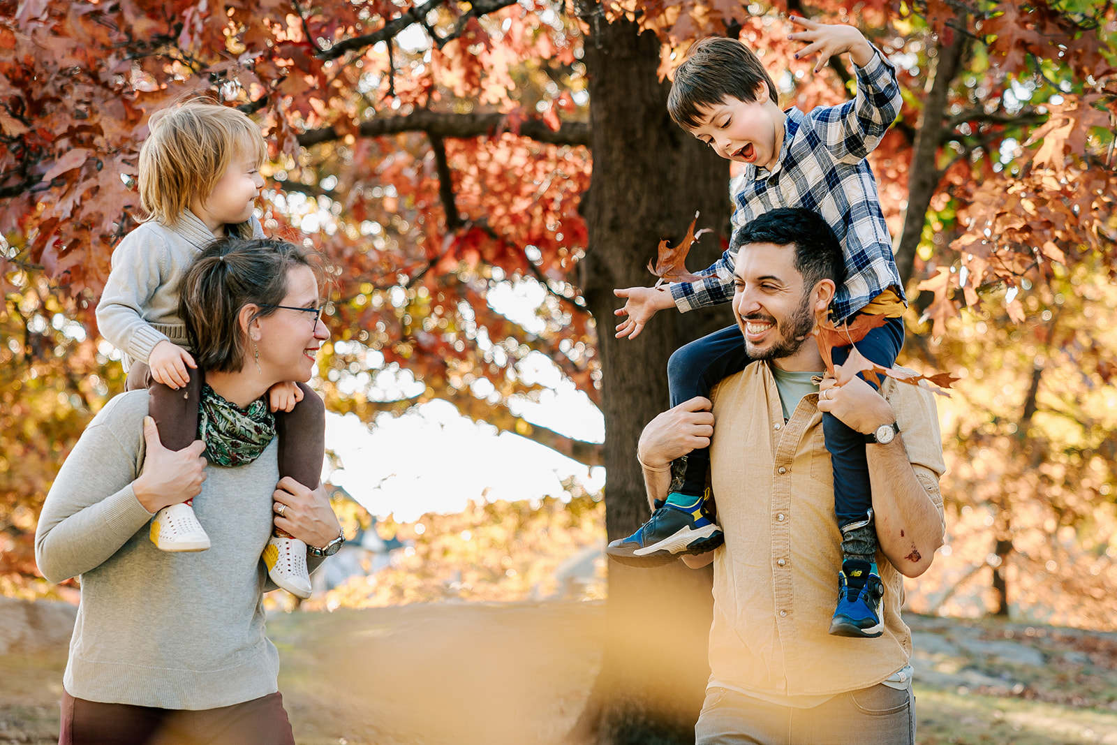 Happy mom and dad play in a park in fall with their toddlers on their shoulders after meeting Boston Nannies