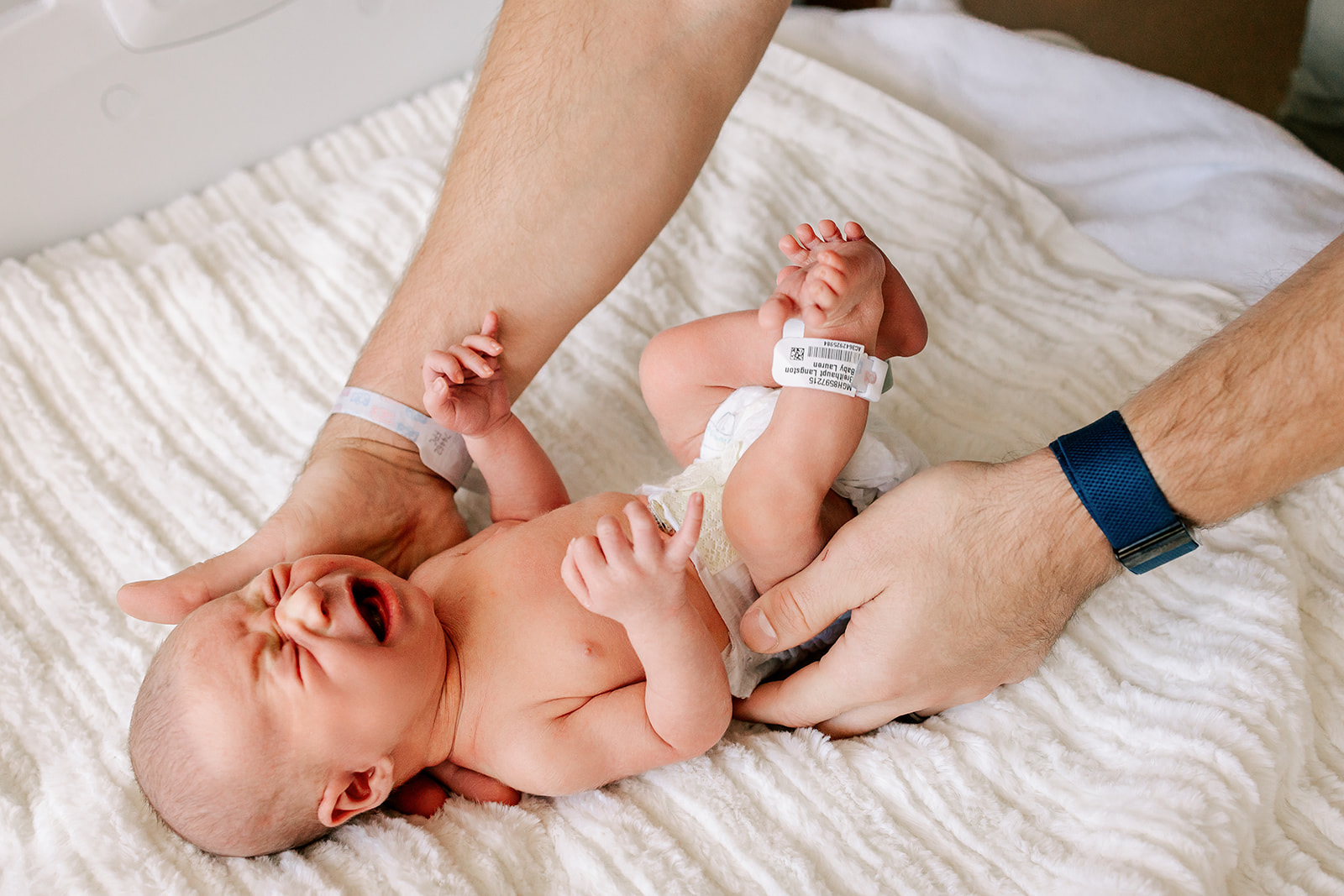 Dad reaches to pick up his crying newborn in the hospital crib while using diaper service in Boston