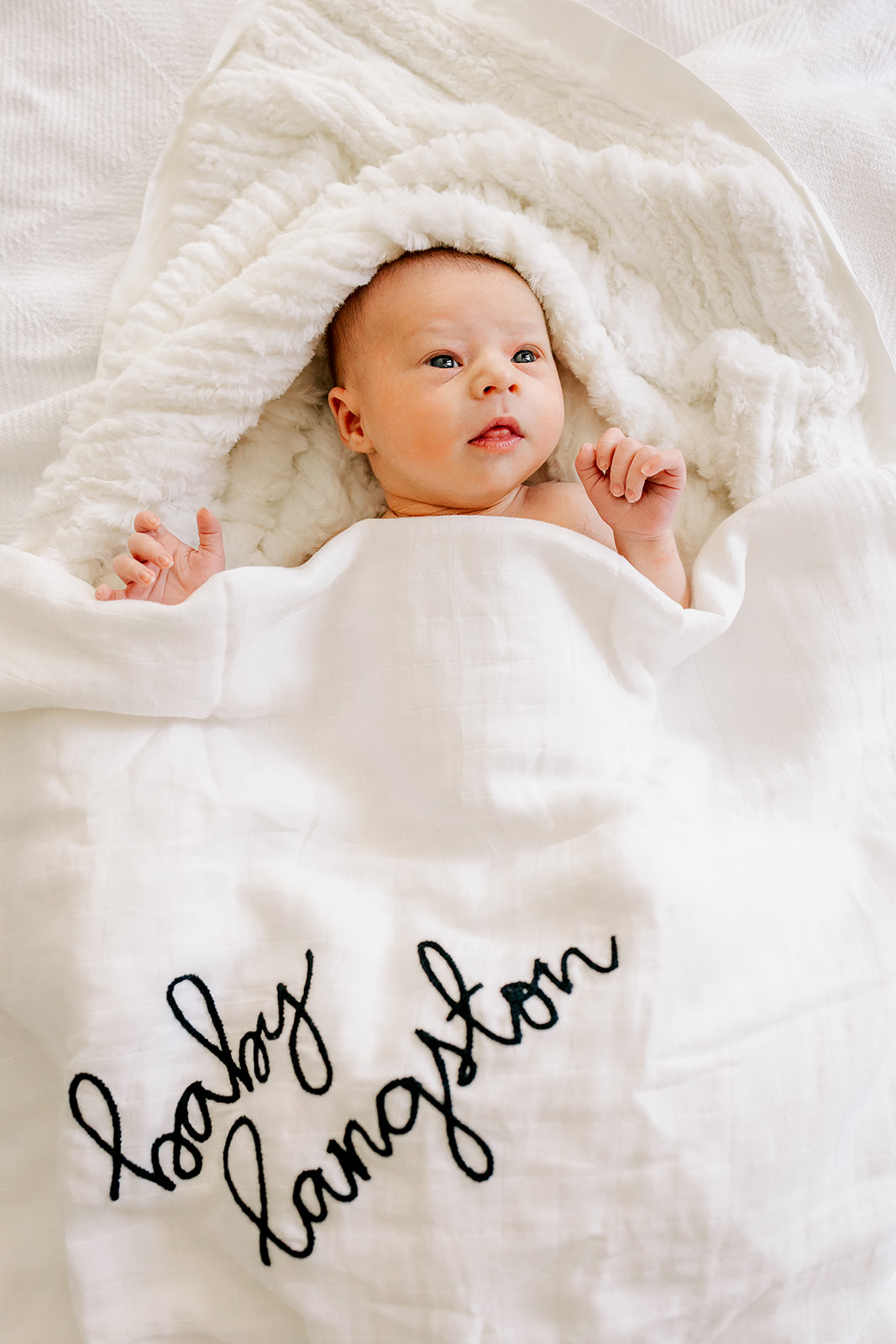 A newborn baby lays in a custom embroidered blanket with eyes open on a bed while using diaper service in Boston