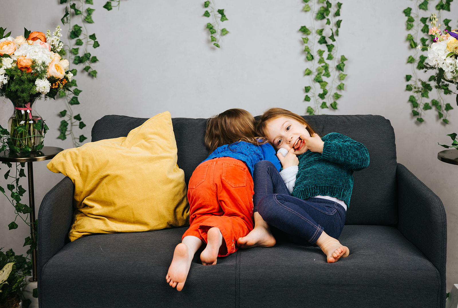 Two toddler girl in orange and blue jeans play and make silly faces on a couch in a studio