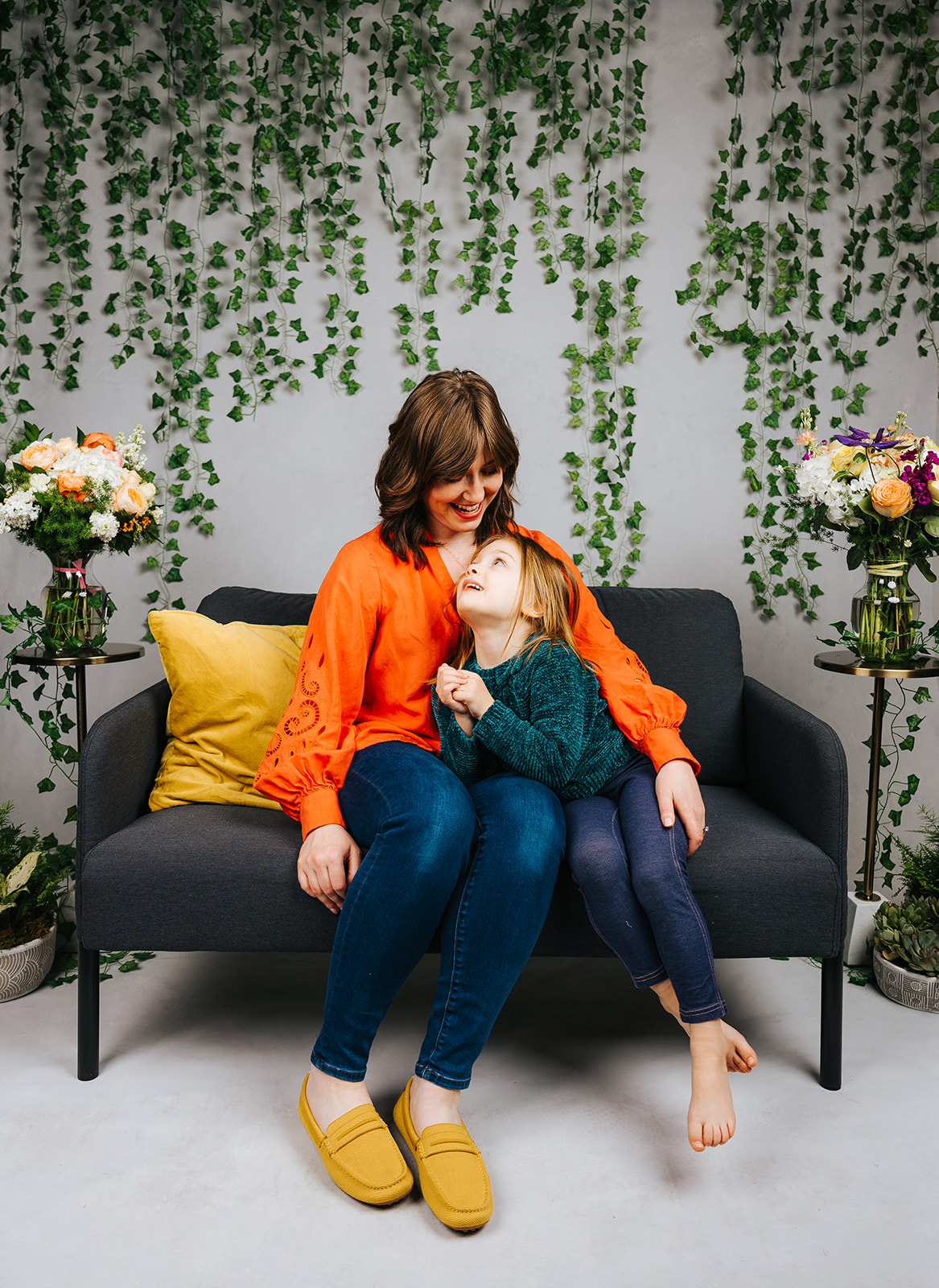 A young girl in a blue sweater cuddles her mom on a couch in a studio before visiting indoor playgrounds in Boston