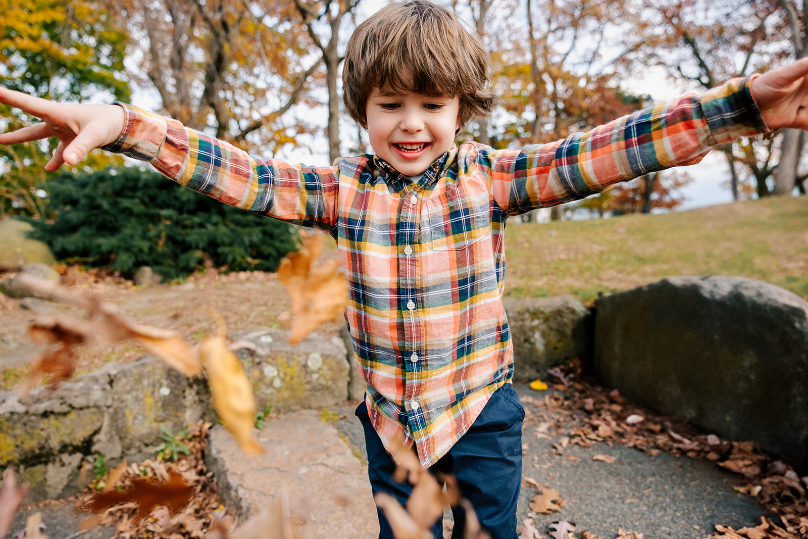 A toddler boy in plaid tosses leaves in the air in a park while laughing